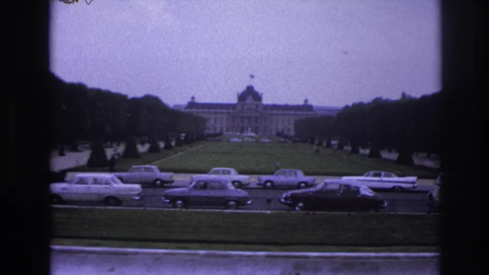 Paris home-movie frame from 1967 showing a formal green approach, clipped trees, a distant civic-looking building, and period boulevard traffic.