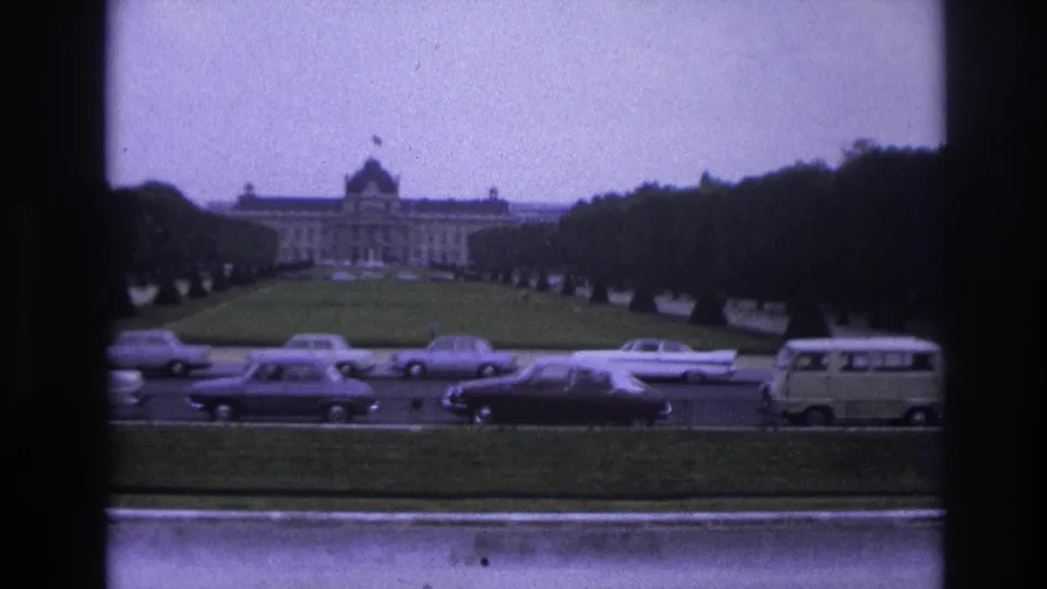 1967 Paris frame showing a formal lawn, rows of trees, several mid-century cars, a light van, and a distant grand building under a purple cast.