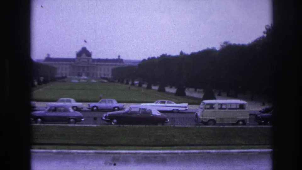 1967 Paris home-movie view of a formal lawn, distant classical building, and mid-century cars passing along the road.
