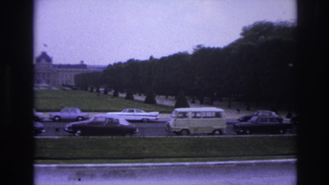 Tree-lined roadway with cars, a light van, and a distant large building behind lawns.