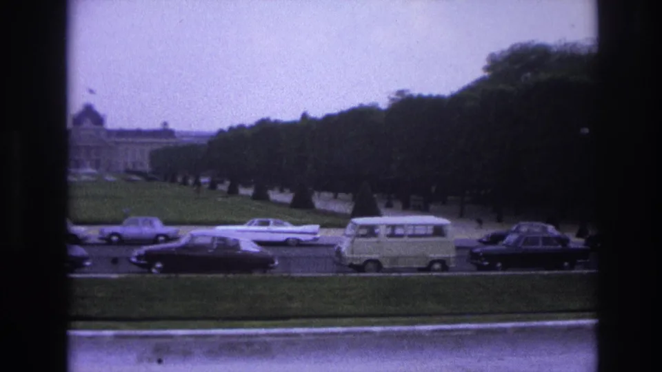 1967 Paris street view framed by dark edges, showing cars and a van moving before a distant grand building and trees.
