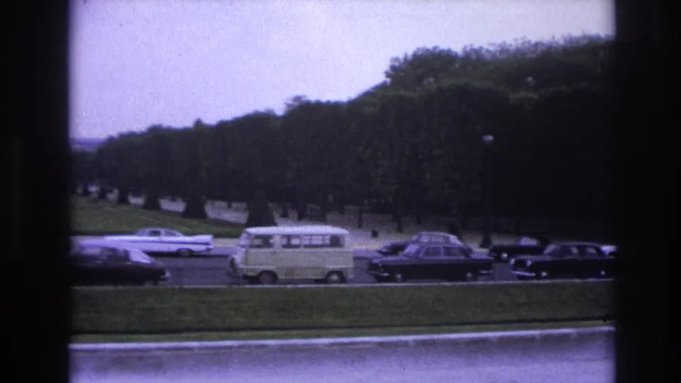 1967 Paris home-movie frame with a light van and dark cars crossing a road below dense trees and formal grounds.
