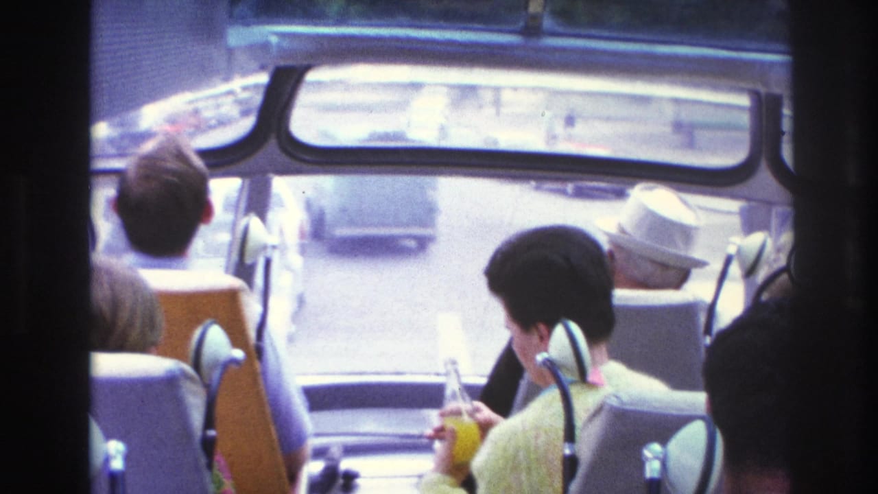 1967 Paris bus interior with seated tourists, including a woman in yellow holding a drink and cars visible ahead.