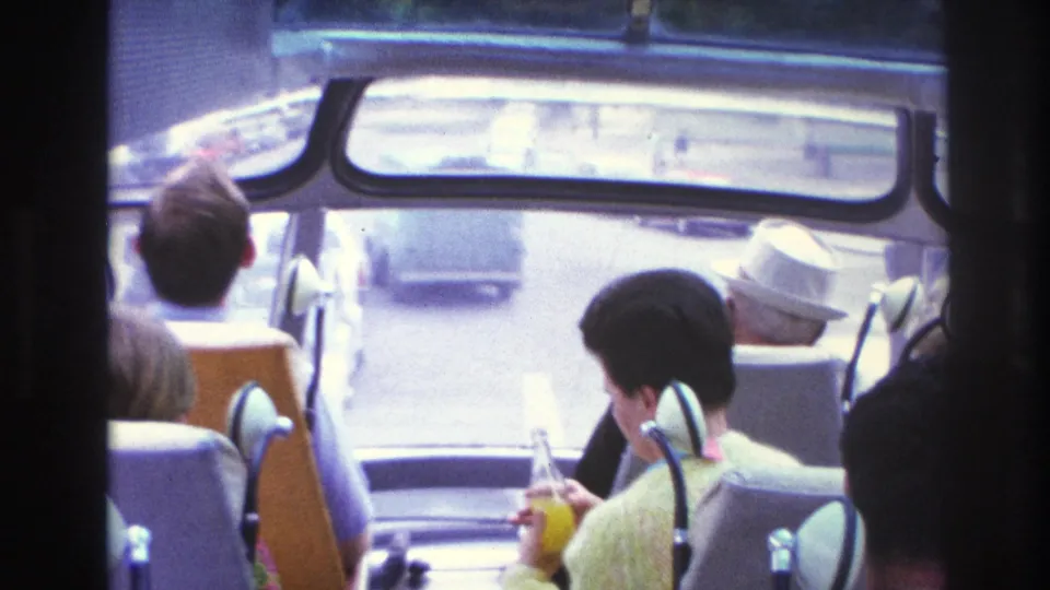 1967 Paris bus interior with seated tourists, including a woman in yellow holding a drink and cars visible ahead.