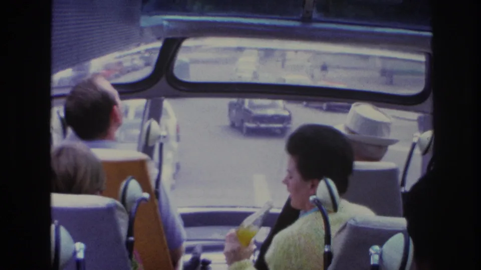 Inside a 1967 Paris tour bus, tourists sit near the front while mid-century cars move along the street outside.