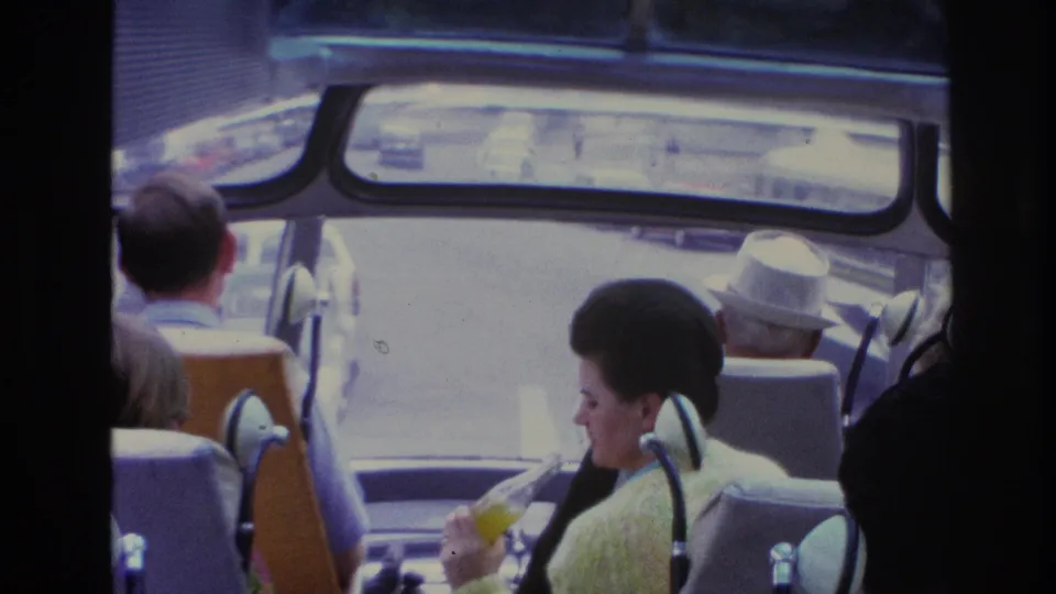 Tourists inside a 1967 Paris bus, with a woman in yellow holding a drink and a man in a white hat facing the front windows.