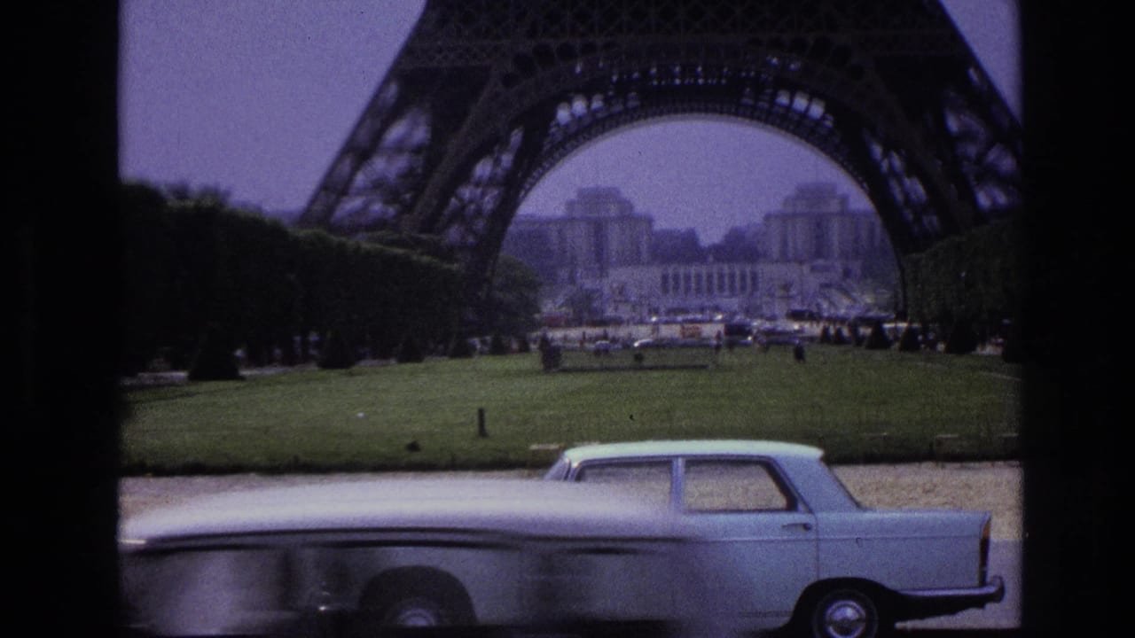 Eiffel Tower base with lawn and a pale vintage sedan crossing the foreground.