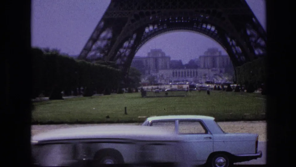 Eiffel Tower seen across the Champ de Mars in Paris, with a pale vintage sedan passing in the foreground.