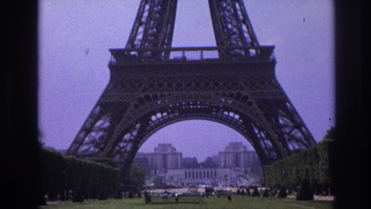 1967 Paris home-movie view of the Eiffel Tower base through a dark frame, with lawns, clipped hedges, distant buildings, and small visitors below.