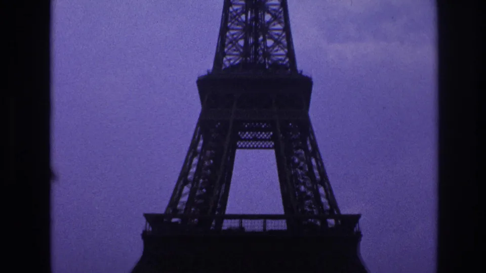 1967 Paris home-movie frame looking upward at the Eiffel Tower midsection, with heavy dark framing and purple-toned overcast sky.