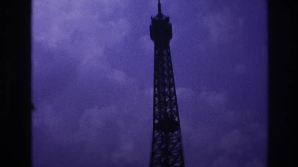 1967 Paris 8mm frame with the Eiffel Tower spire and upper lattice centered against a muted cloudy sky.