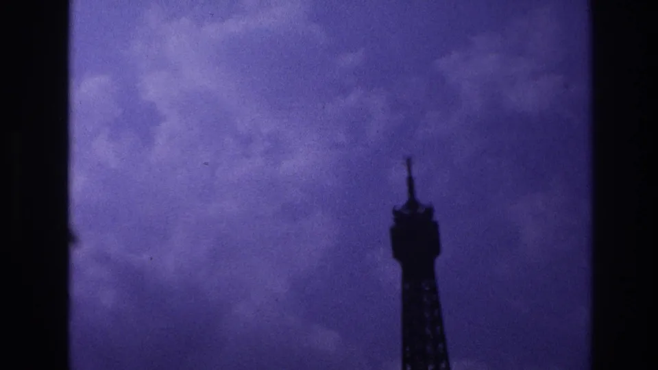 1967 Paris home-movie frame with the Eiffel Tower top silhouetted at right under a purple, overcast sky.