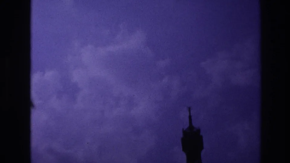 1967 Paris home-movie frame showing the Eiffel Tower top small at lower right beneath heavy purple clouds.