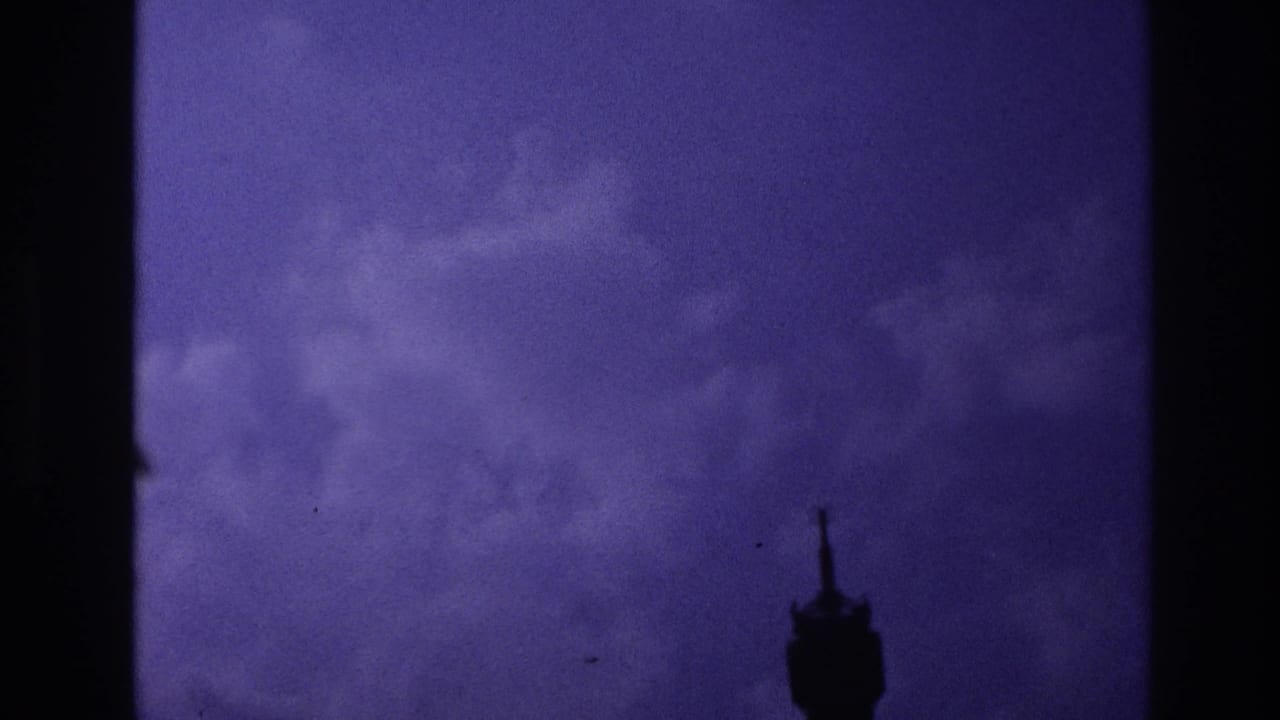 Underexposed 8mm frame from the Paris transit sequence, showing a dark interior silhouette against blue-purple window light.