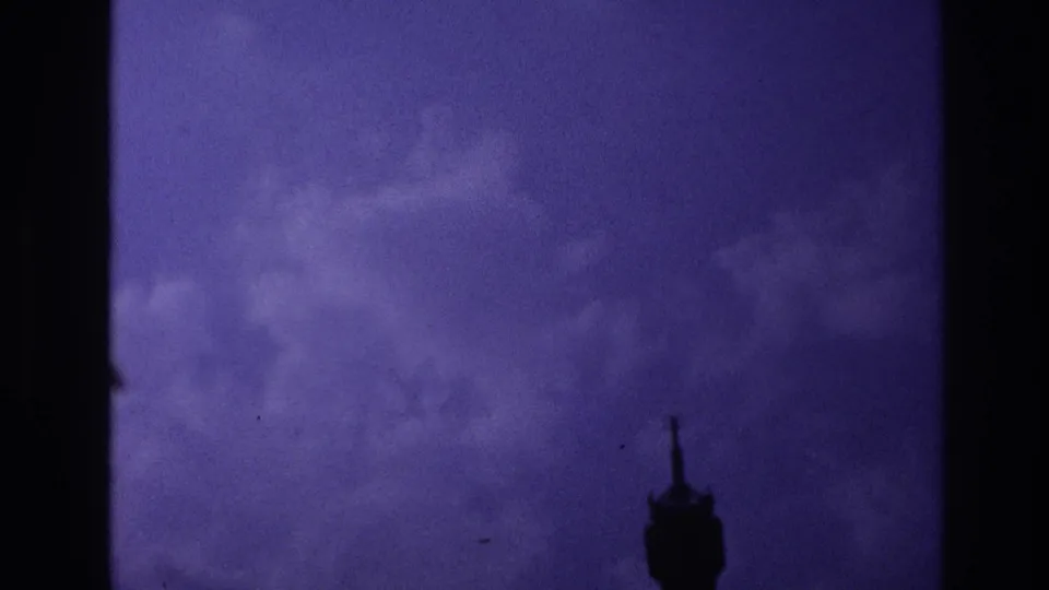 Underexposed 8mm frame from the Paris transit sequence, showing a dark interior silhouette against blue-purple window light.