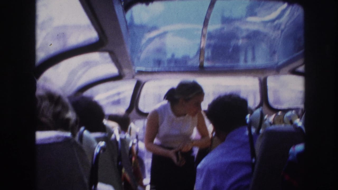1967 Paris home-movie frame showing a woman in a sleeveless top leaning in the aisle among passengers under a curved glass roof.