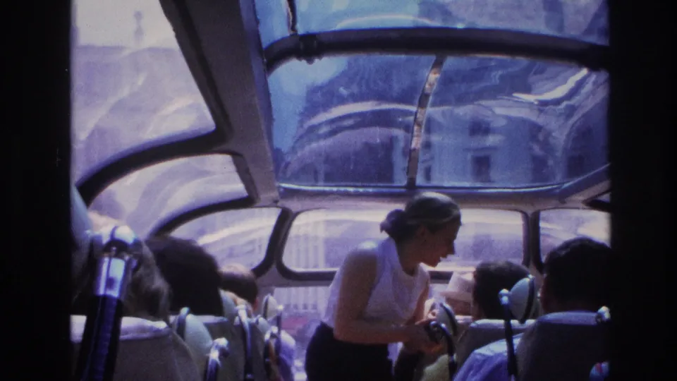 1967 Paris home-movie frame of passengers in a glass-roofed transit vehicle, with a woman standing near the aisle and city light beyond the windows.