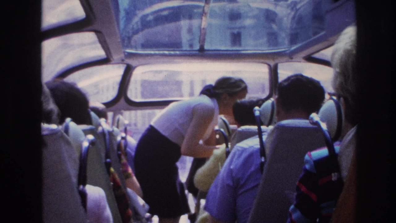 Woman leans over near a seated passenger while tourists face the windows of a bus.