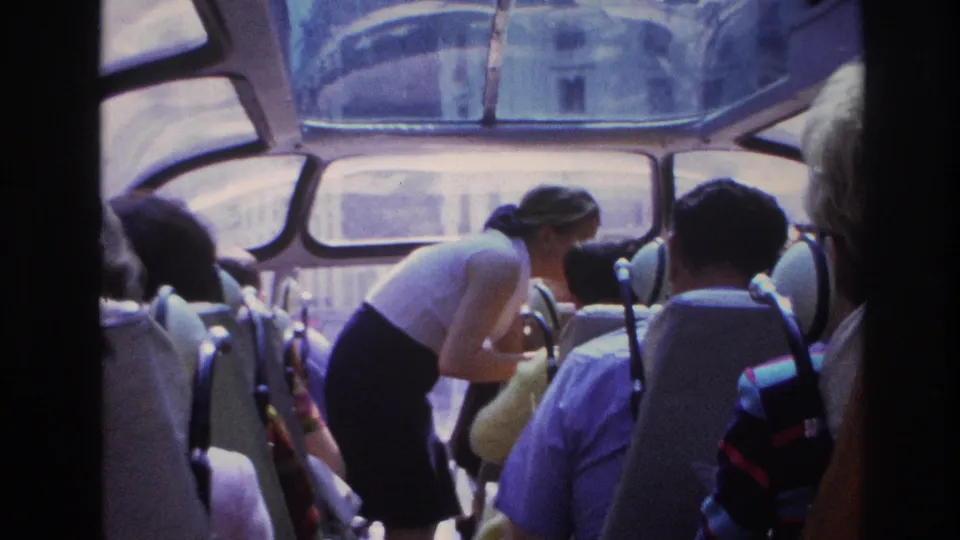 On a Paris sightseeing bus in 1967, a woman bends beside seated passengers as buildings appear through the curved front windows.