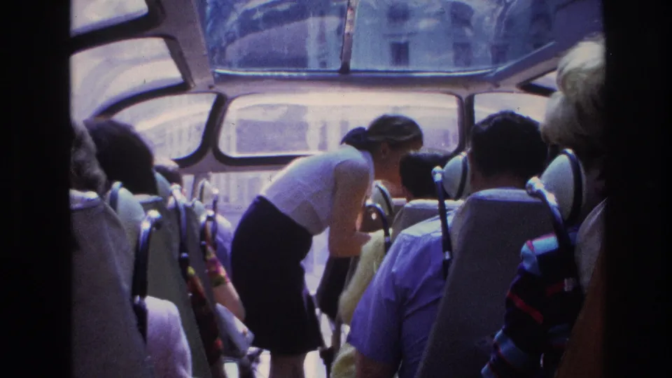1967 Paris home-movie frame showing tourists seated inside a bus while a woman in a white top leans toward a passenger.