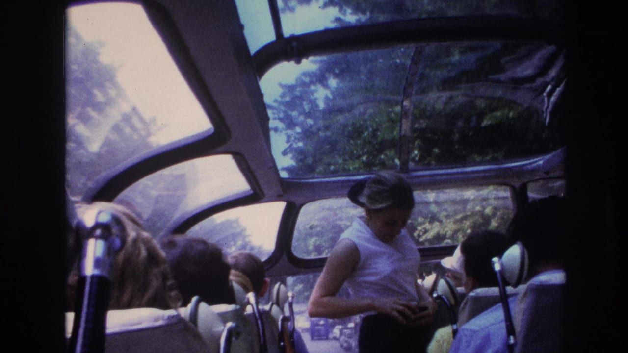 1967 Paris tour bus interior with seated passengers, a standing female guide, curved windows, and trees visible through the glass roof.