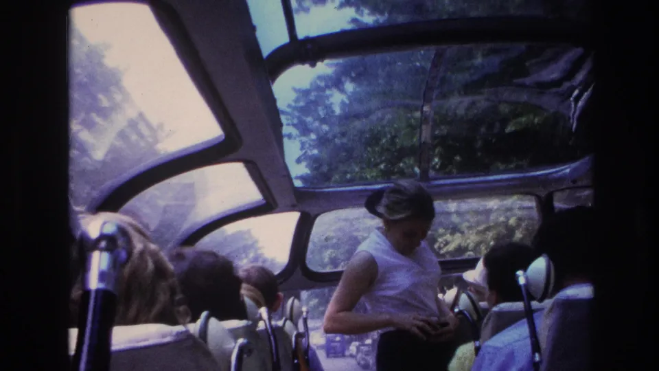 1967 Paris tour bus interior with seated passengers, a standing female guide, curved windows, and trees visible through the glass roof.