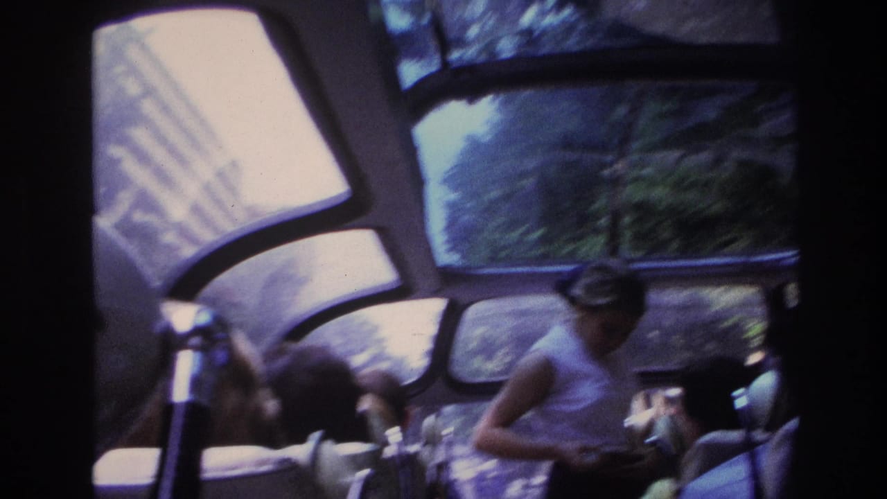 A woman stands in the aisle of a tour bus as passengers sit by large windows.