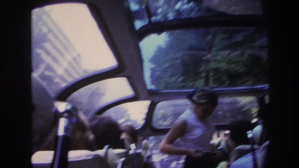 Inside the 1967 Paris sightseeing bus, the guide stands near seated tourists with trees and bright reflections visible through the windows.