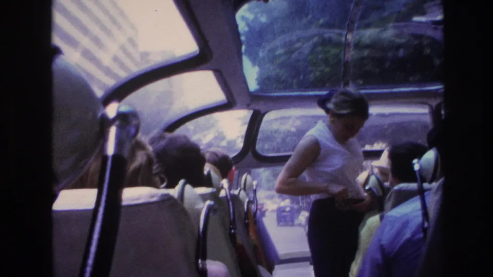 A handheld 8mm view inside a Paris tour coach shows the guide beside seated passengers under the curved glass ceiling.