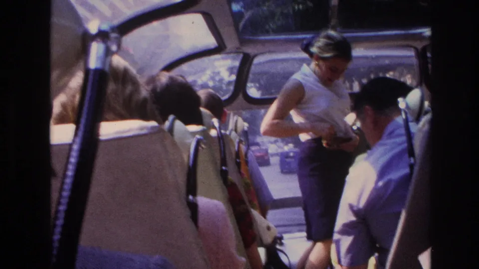 The Paris tour bus interior shows the guide near the front, seated tourists, a uniformed man, and greenery visible beyond the glass windows.