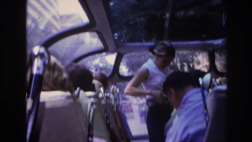 1967 Paris home-movie frame showing bus passengers, curved windows, and a woman in a white top near the aisle.
