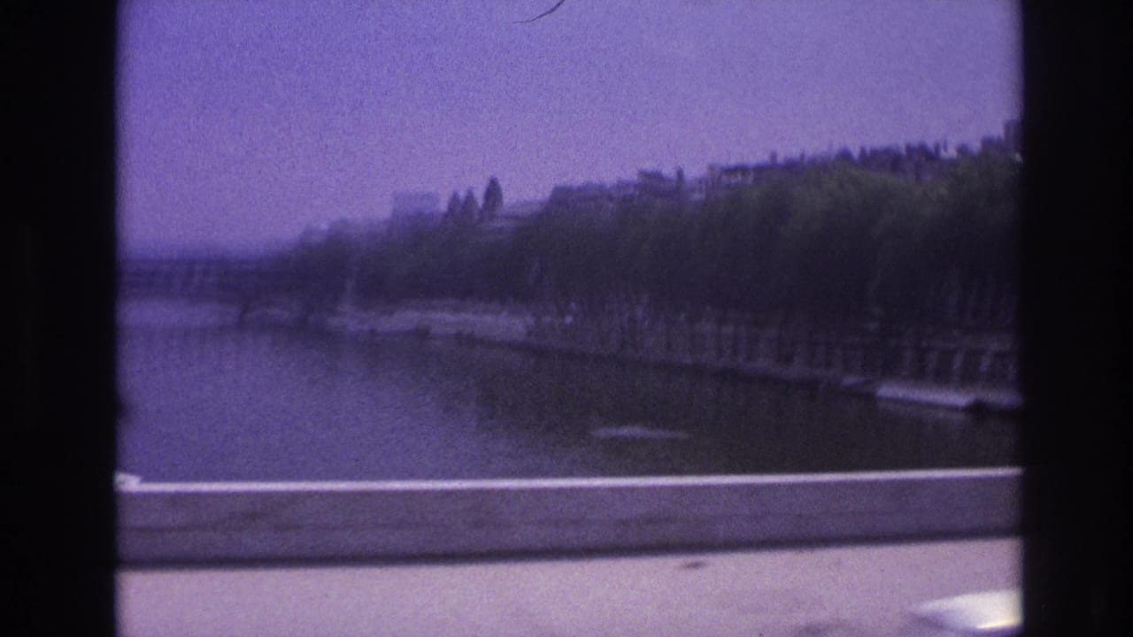 1967 Paris home-movie frame looking past vehicle window bars toward a tree-lined waterway, embankment, and road barrier.