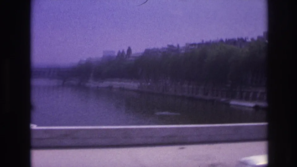 1967 Paris home-movie frame looking past vehicle window bars toward a tree-lined waterway, embankment, and road barrier.