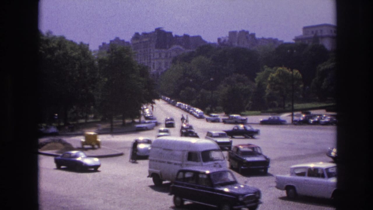 White van and dark sedans travel across a wide roadway with parked cars and trees beyond.