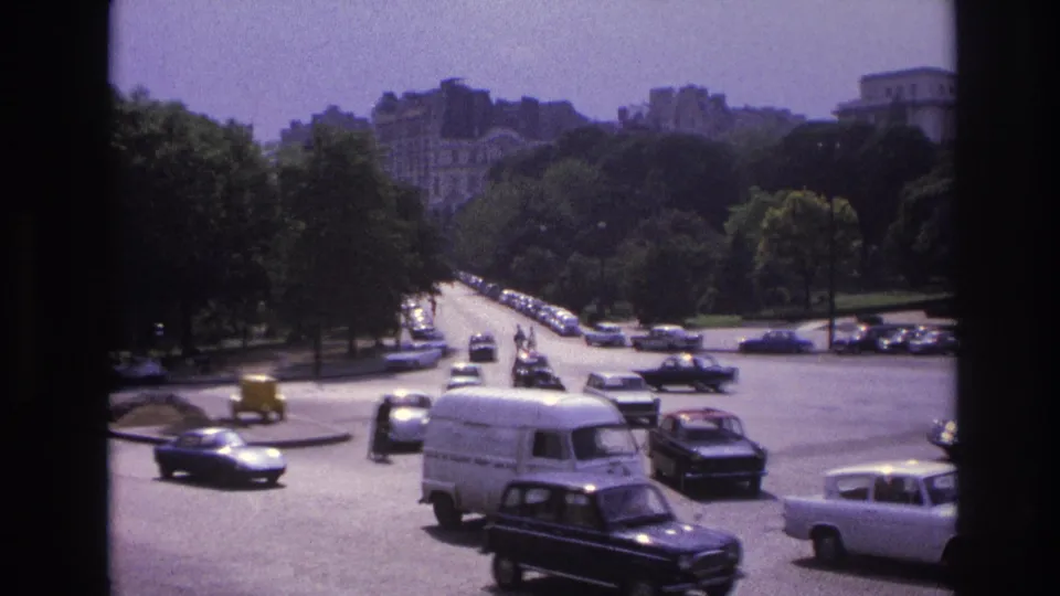 Wide Paris traffic scene with a white van, dark sedans, parked cars, trees, and distant buildings.