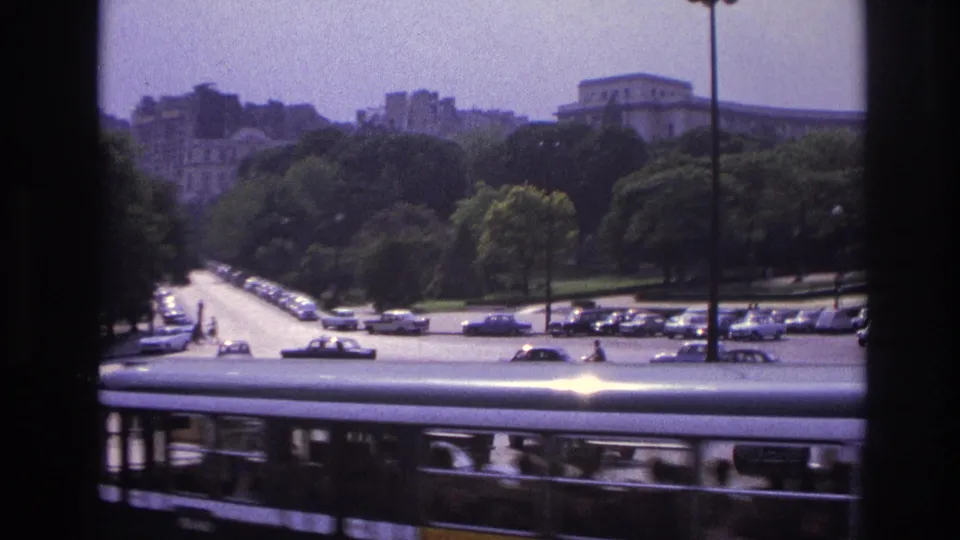 A bus crosses the foreground of a 1967 Paris traffic view, with cars, trees, and distant buildings behind it.
