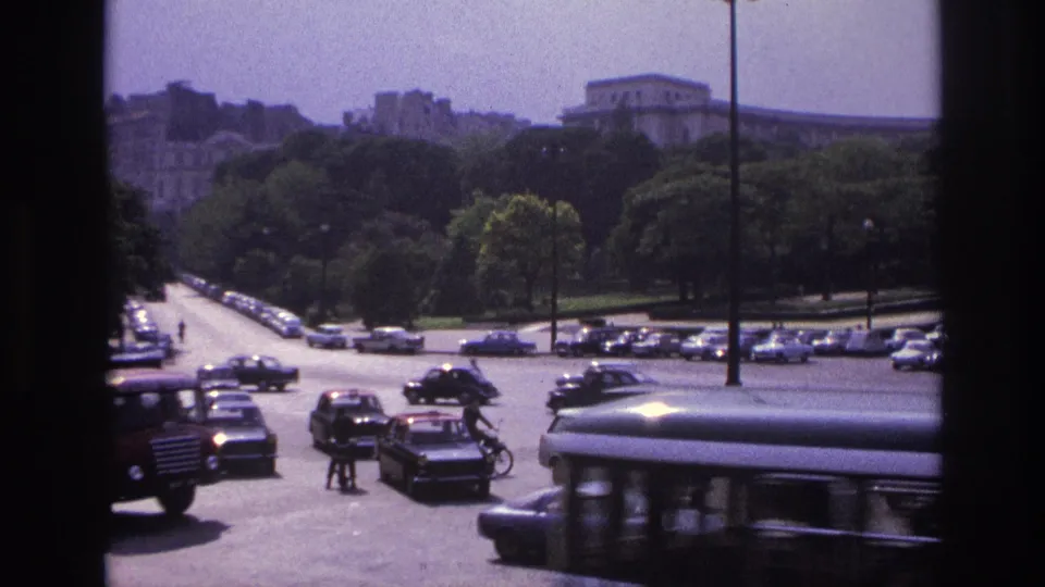 1967 Paris street view with moving and parked period cars, pedestrians, trees, and a large building beyond the roadway.