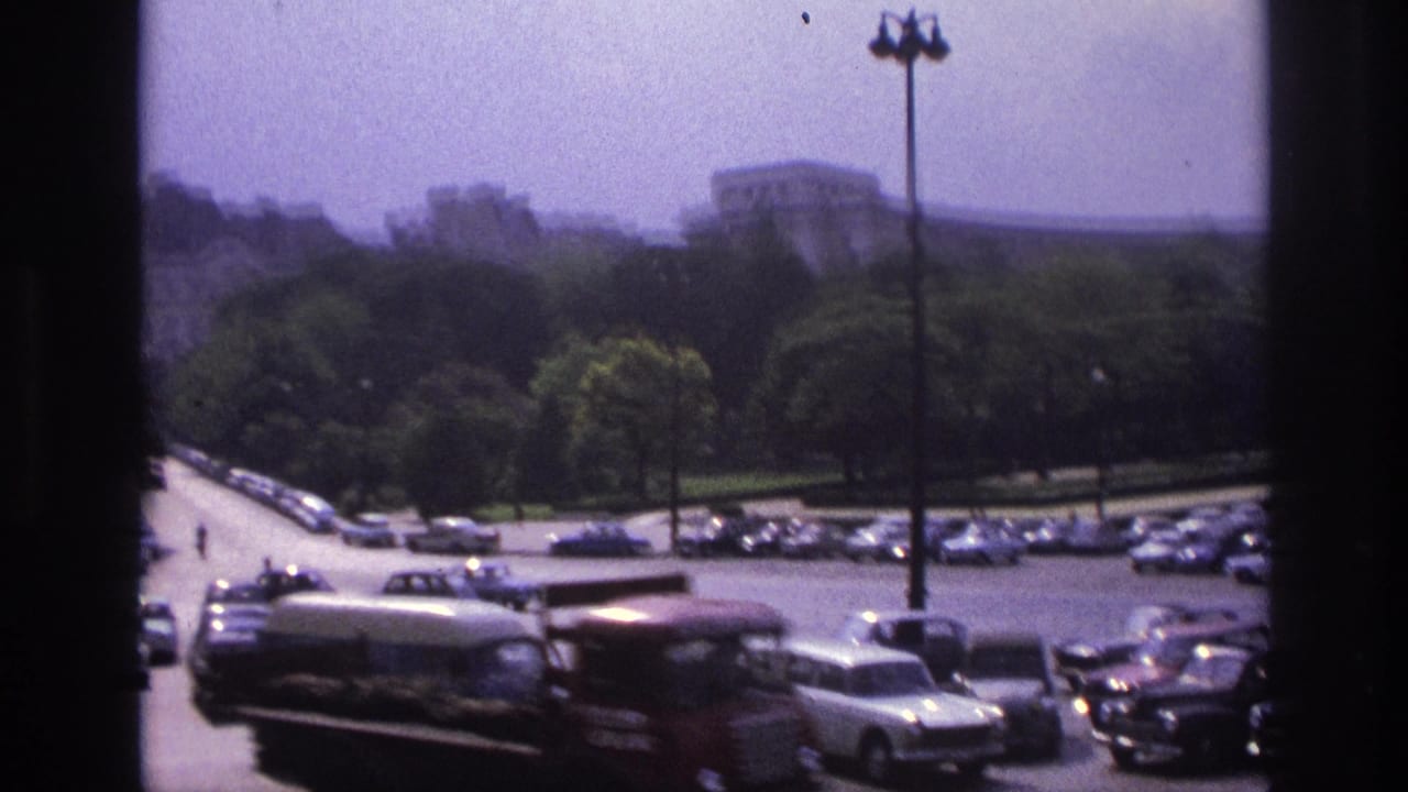Traffic passes a parking area with a red truck and many parked cars.