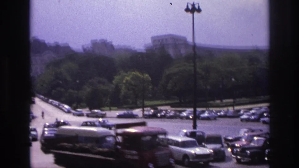 1967 Paris traffic scene with a red delivery-style truck, station wagon, parked cars, trees, and a large building in the background.