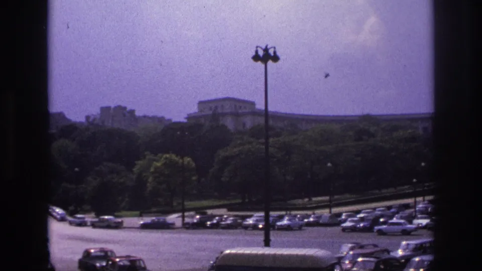 1967 Paris street view showing rows of parked cars, moving vehicles, a tall street lamp, trees, and distant buildings.