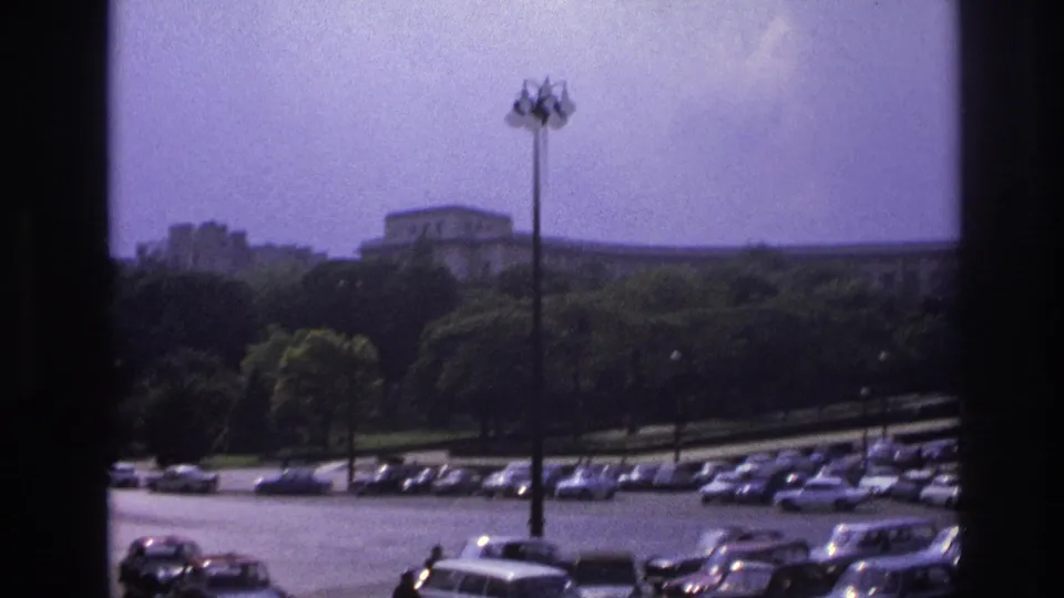 1967 Paris roadway with parked period cars, passing vehicles, tall lamps, trees, and a large building beyond the park-like grounds.