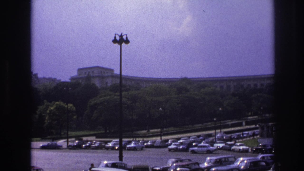 1967 Paris home-movie frame showing parked mid-century cars below a tree line and long formal building, seen through dark window edges.