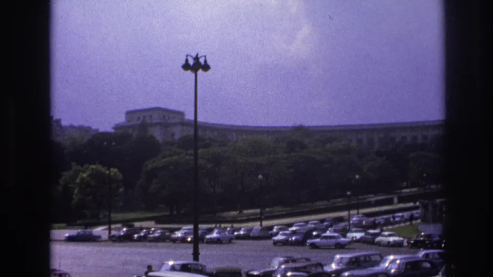 1967 Paris home-movie frame showing parked mid-century cars below a tree line and long formal building, seen through dark window edges.