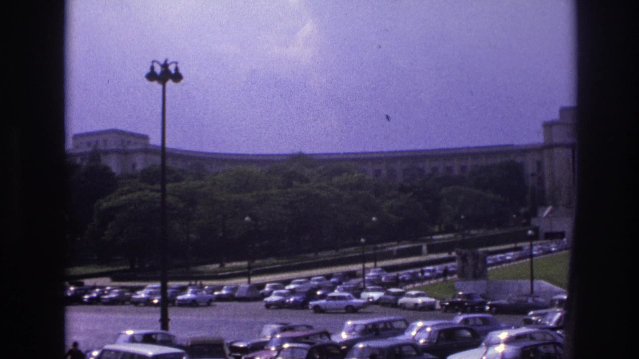 Wide parking area with many cars, a lamppost, trees, and a long building in the background.