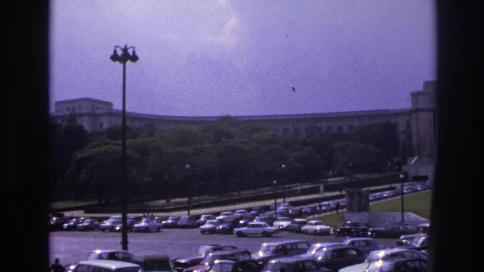 1967 Paris frame of a large outdoor parking area with rows of cars and a long building behind trees, bordered by dark interior framing.