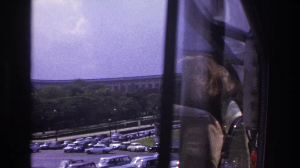 1967 Paris frame showing rows of parked cars and a distant building, with a partial passenger figure visible beside the window frame.