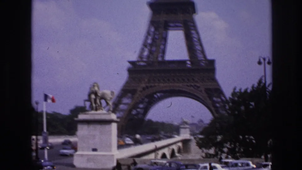 1967 Paris frame with the Eiffel Tower rising behind an equestrian statue, French flag, bridge, and moving cars.