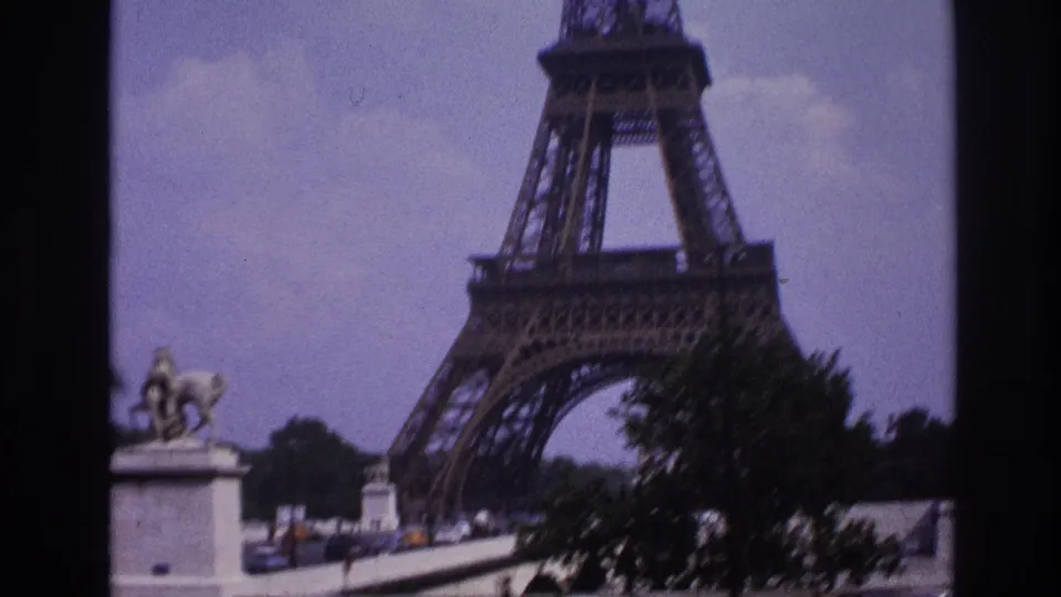 1967 Paris home-movie view of the Eiffel Tower base beside a bridge, statue, trees, and passing cars.