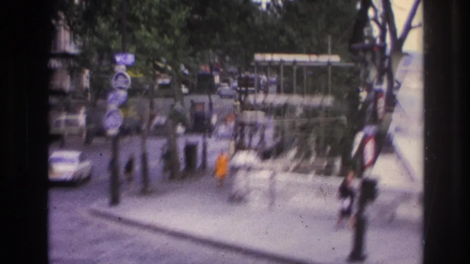 1967 Paris home-movie frame showing a nearby street corner with pedestrians, road signs, cars, trees, and a kiosk-like structure.