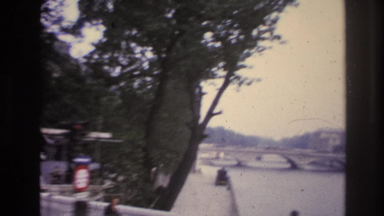 1967 Paris home-movie frame looking past trees and a riverside wall toward an arched bridge over the river.